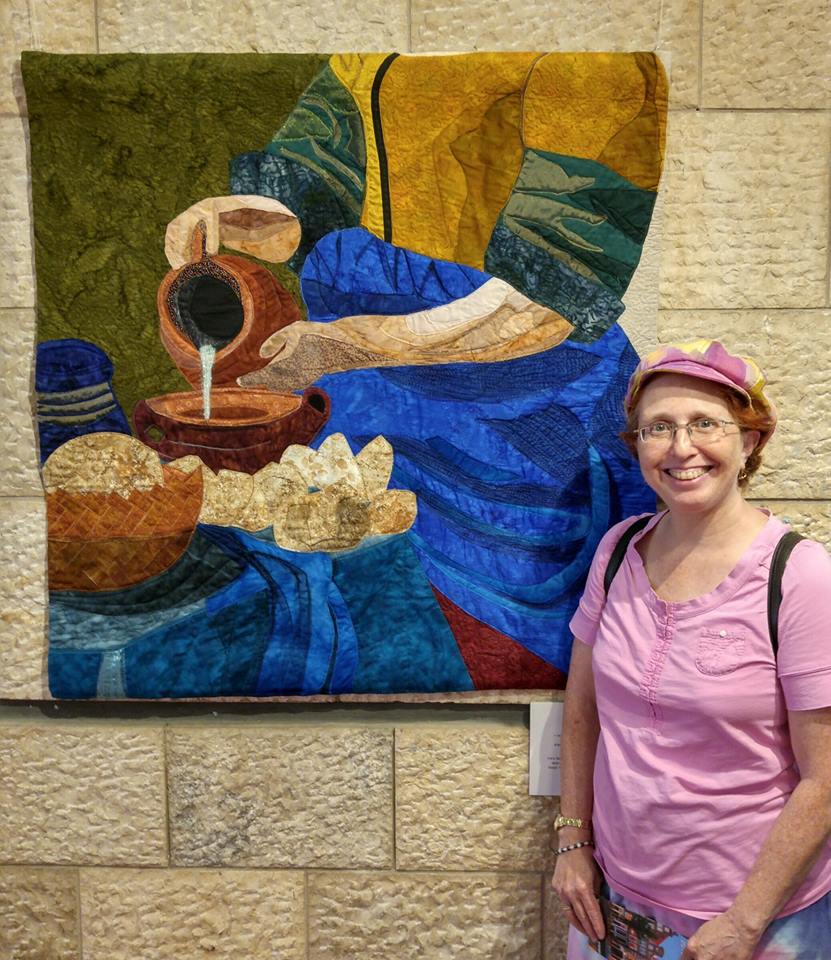 Israeli quiltmaker stands in front of her art piece depicting a woman pouring milk out of a jug